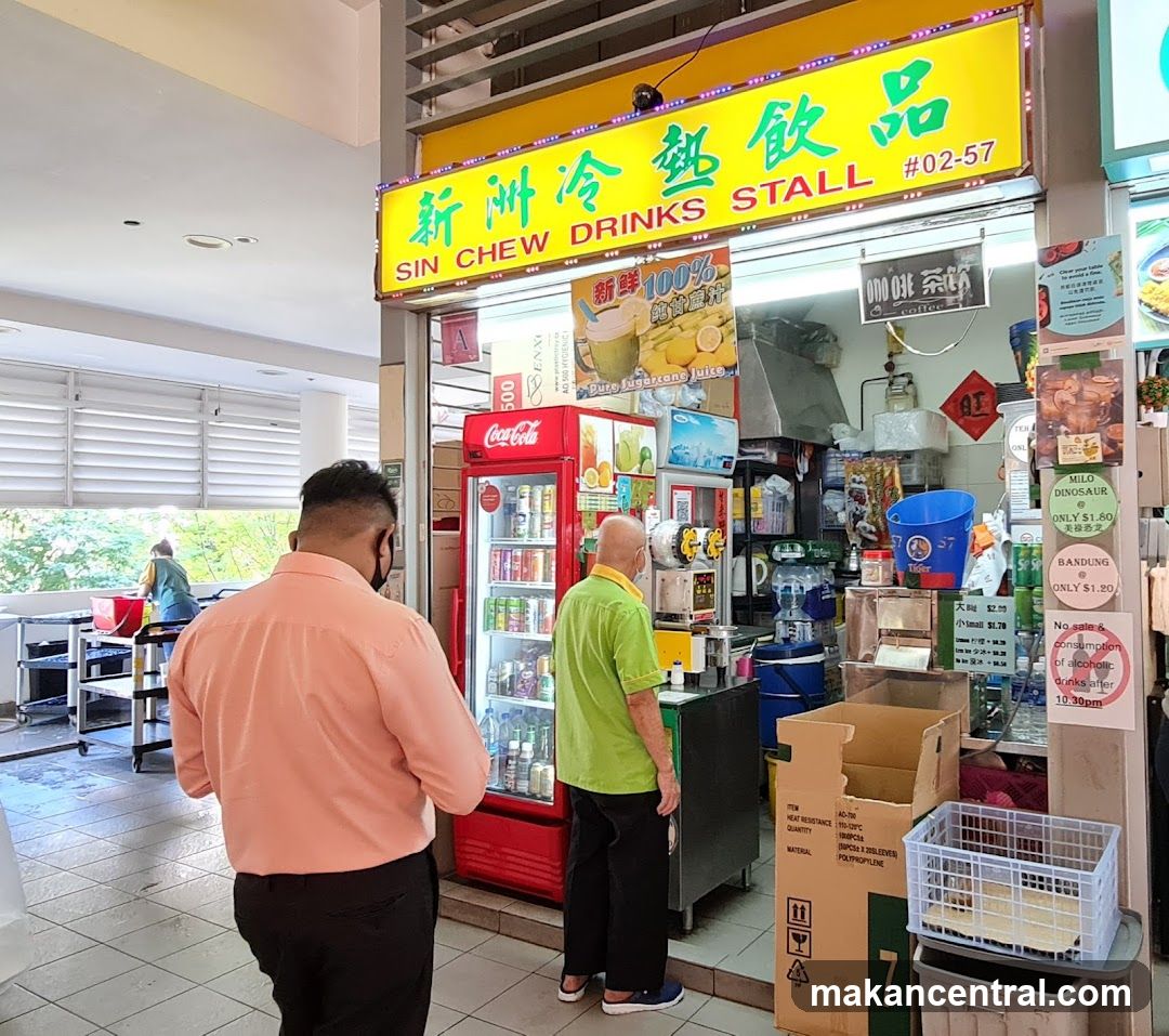 A drinks stall with a bright yellow sign that reads "SIN CHEW DRINKS STALL #02-57" in English and Chinese. A red Coca-Cola refrigerator is visible, stocked with various beverages. Two customers are present, one in a peach shirt and another in a green shirt. Various boxes and supplies are stacked around the stall.