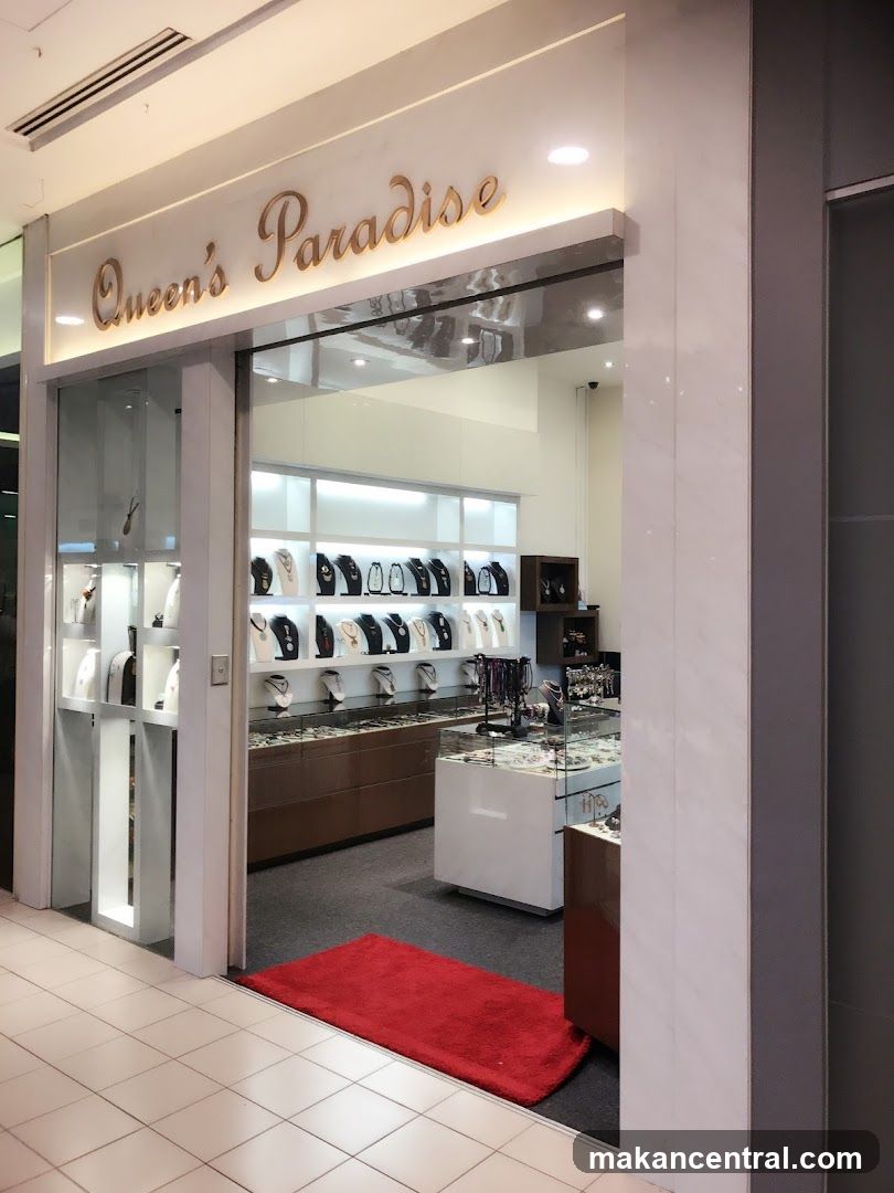 The exterior of a jewelry store called Queen's Paradise. The store has a clean, modern facade with large glass windows and a prominent sign above the entrance. Inside, displays of necklaces and other jewelry are visible. A red rug is placed just inside the entrance.