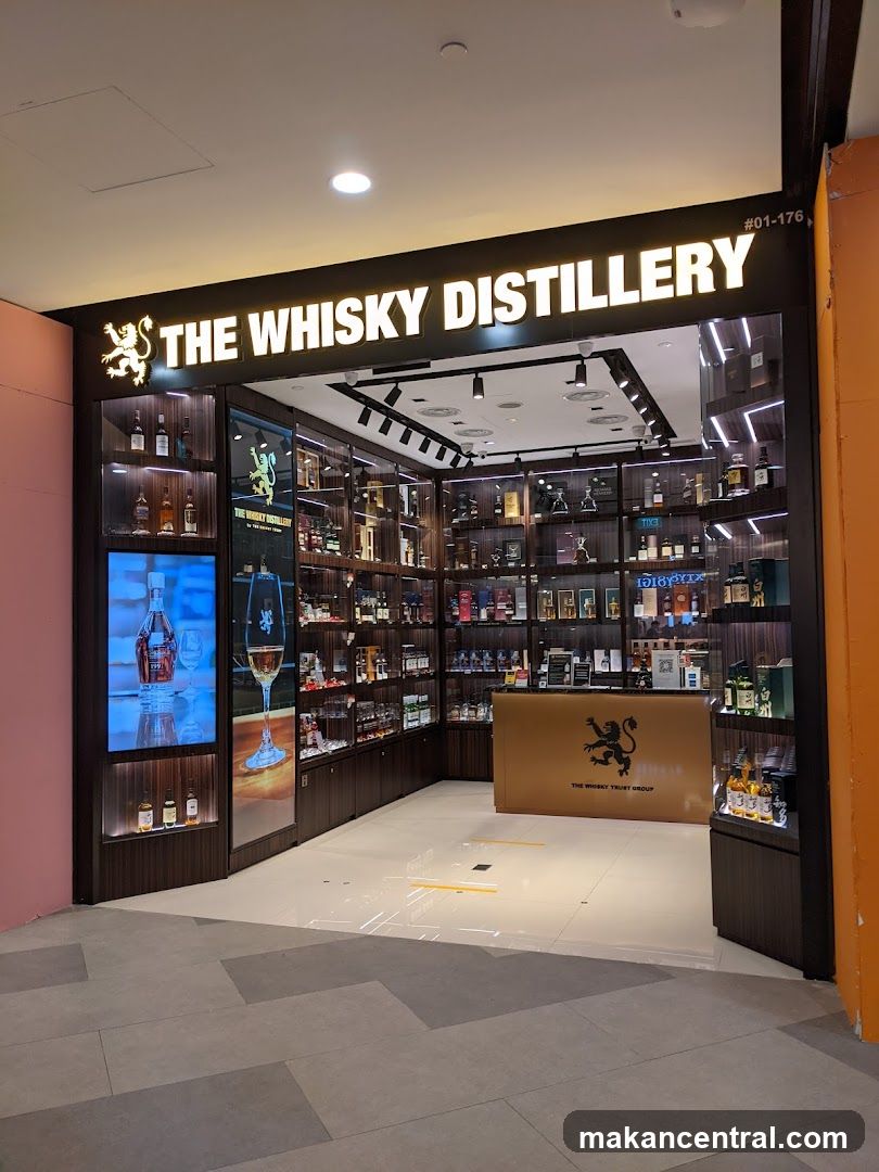 A well-lit storefront for a whisky shop. The exterior features a prominent illuminated sign that reads "THE WHISKY DISTILLERY" with a lion logo. Inside, shelves are stocked with numerous bottles of whisky. A counter with the "THE WHISKY TRUST GROUP" logo is visible. Digital displays showcase whisky bottles and a glass.
