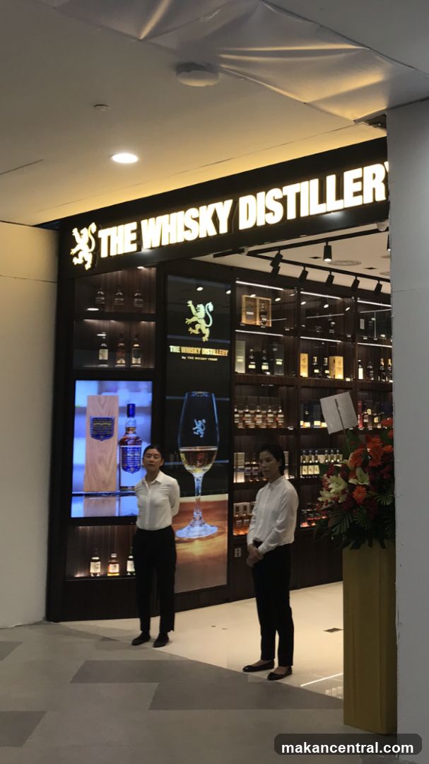 The exterior of a shop called "The Whisky Distillery" is shown. The shop has a large illuminated sign with the name and a lion logo. Inside, shelves are stocked with various bottles of whisky. Two women in white shirts and black pants stand in front of the shop. A large display features a bottle of whisky and a glass.