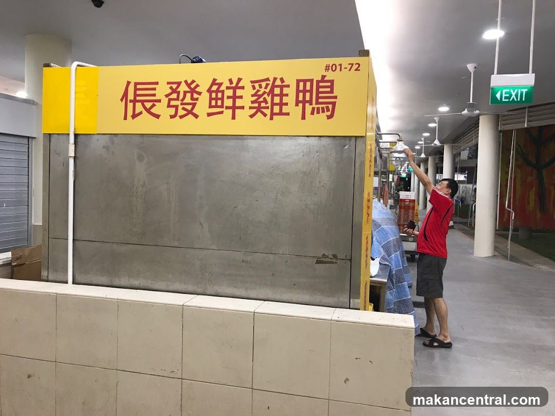 A man in a red shirt and grey shorts reaches up towards a yellow sign with red Chinese characters and the number #01-72. The sign appears to be above a stall selling fresh chicken and duck. The stall has a concrete facade. In the background, an "EXIT" sign is visible.