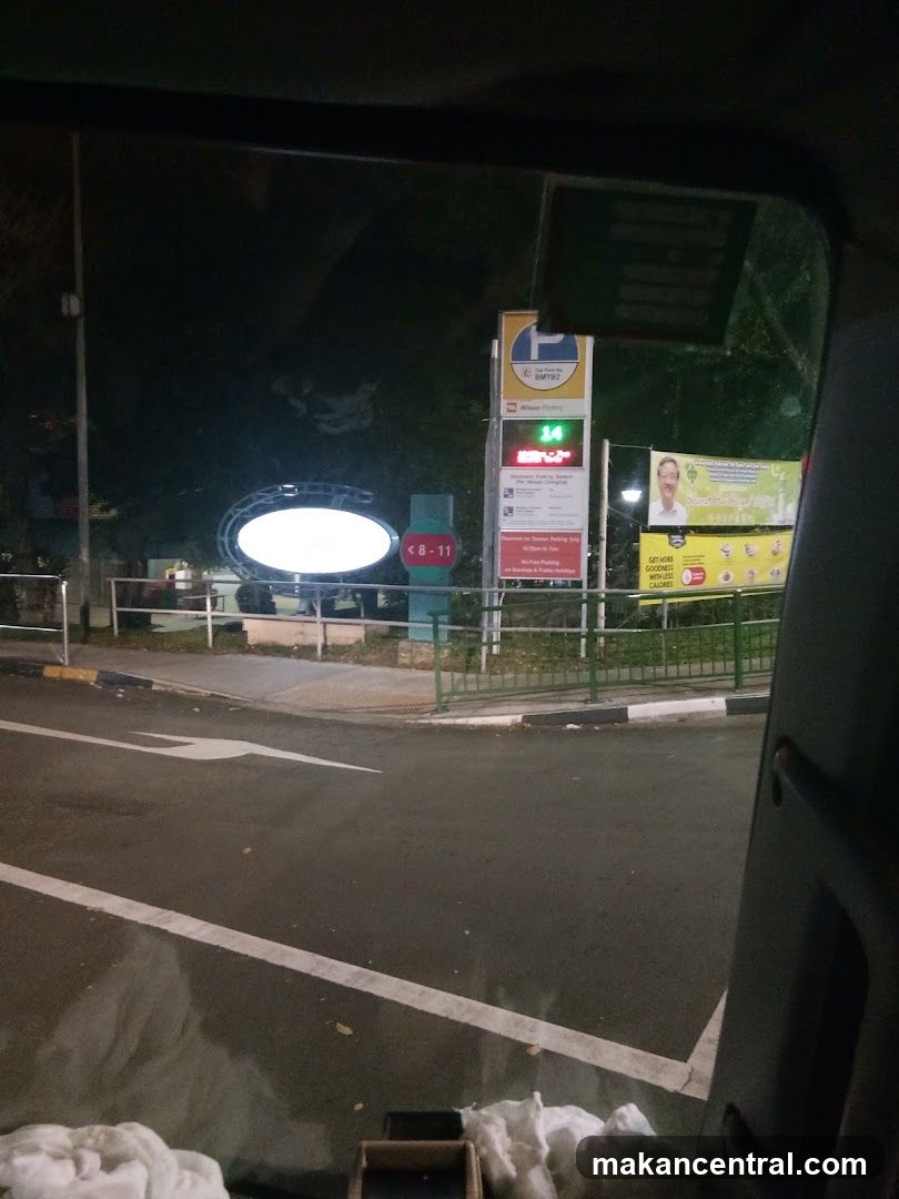 A night-time view of a parking lot with various signs. One sign indicates "Car Park No. BMTB2 Wilson Parking" and displays a green number "14" indicating available lots. Another sign shows a man's face and promotional text. An oval-shaped illuminated sign is also visible.