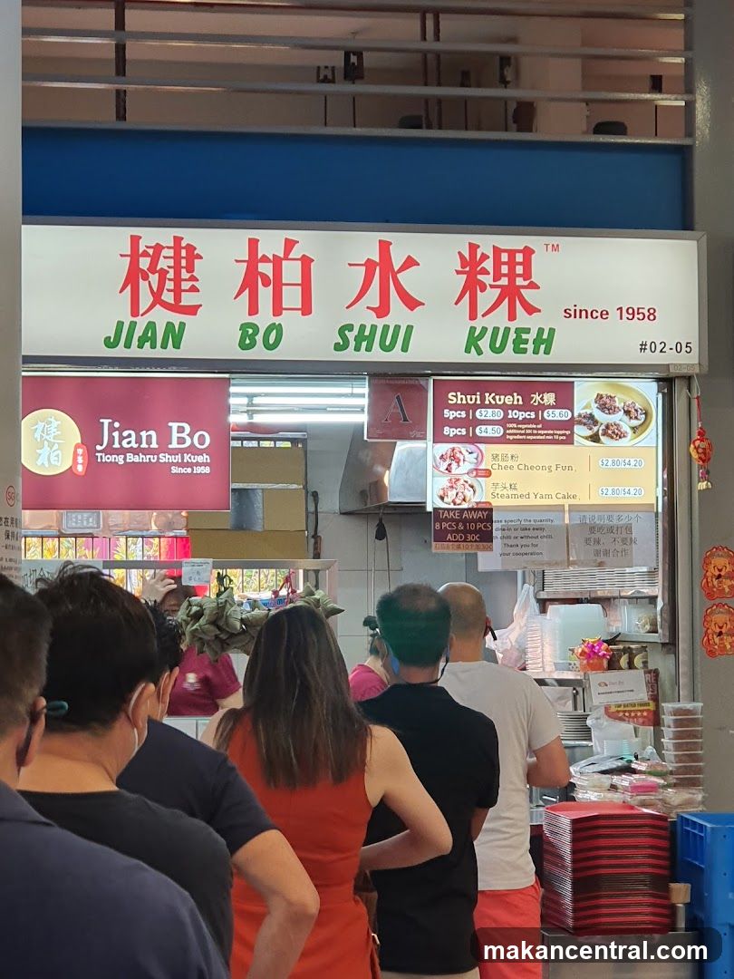 A queue of people waiting outside a food stall. The stall has a large sign with Chinese characters and the name "Jian Bo Shui Kueh" in English, along with "since 1958". Below the main sign, there is a smaller sign with the stall's name and logo. A menu board is visible to the right, displaying prices for Shui Kueh, Chee Cheong Fun, and Steamed Yam Cake.