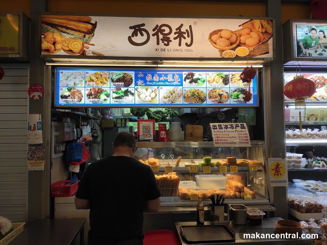 A food stall display with various dishes shown on a backlit menu board. A man is standing in front of the counter, facing away from the camera. The stall has a sign with Chinese characters and "XI DE LI SINCE 1920s". Below the main menu, there is a sign advertising frozen products like dumplings and wontons. The display case contains an assortment of fried and steamed food items. A "VEGETARIAN" sign is visible on the right.