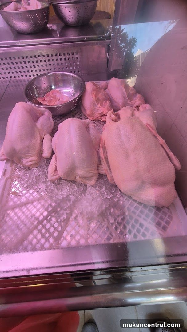 Several whole, raw chickens are displayed on a bed of crushed ice in a refrigerated display case. Above the chickens, metal colanders hold smaller portions of raw poultry. The lighting in the display case casts a pinkish hue on the meat.