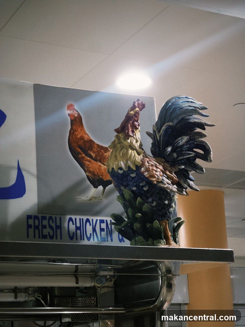 A decorative rooster statue stands in front of a sign that reads "FRESH CHICKEN &" with an image of two chickens behind it. The statue is colorful with blue, green, yellow, and red accents. The sign is blue text on a white background. The image appears to be taken indoors, possibly in a restaurant or food establishment.