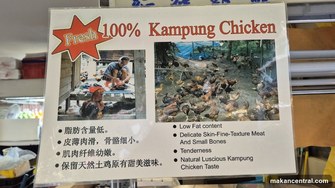 A sign advertising "Fresh 100% Kampung Chicken" with two images. The left image shows a man preparing chicken. The right image shows a flock of chickens in an outdoor enclosure. The sign lists the benefits of the chicken in both Chinese and English, including low fat content, delicate texture, tenderness, and natural taste.