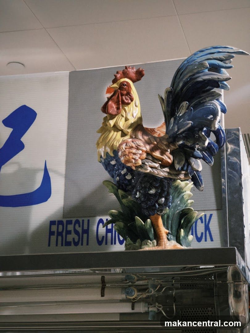 A colorful ceramic rooster statue is displayed above a sign that reads "FRESH CHICKEN". The rooster is detailed with blue, yellow, red, and brown colors, and is perched on green foliage. The sign is in blue lettering against a light gray background. Below the sign, there is a metallic surface with visible wiring and lights.