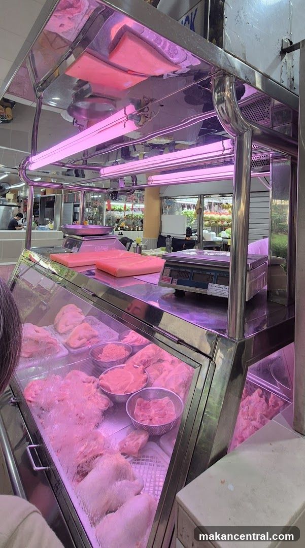 A butcher display case filled with various cuts of raw meat, including whole chickens and smaller portions in bowls. The display is illuminated by pink fluorescent lights, giving the meat a pinkish hue. Scales and pink cutting boards are visible on the counter above the display. In the background, other market stalls with produce can be seen.