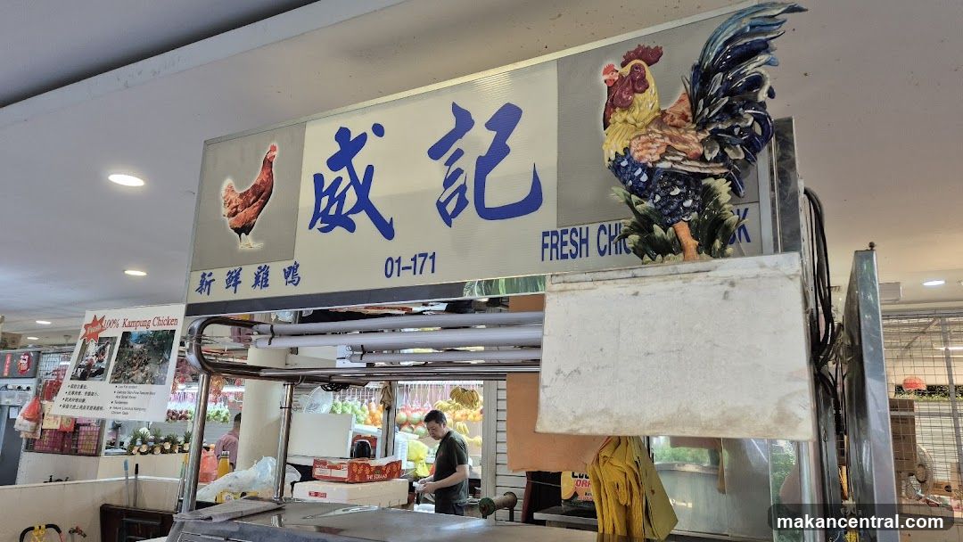 A sign for a fresh chicken stall features Chinese characters, the number 01-171, and the words "FRESH CHICKEN". A decorative ceramic rooster is mounted on the right side of the sign, and an image of a hen is on the left. Below the main sign, a smaller sign advertises "100% Kampung Chicken" with images and bullet points describing its qualities. A man is visible working behind the counter in the background.