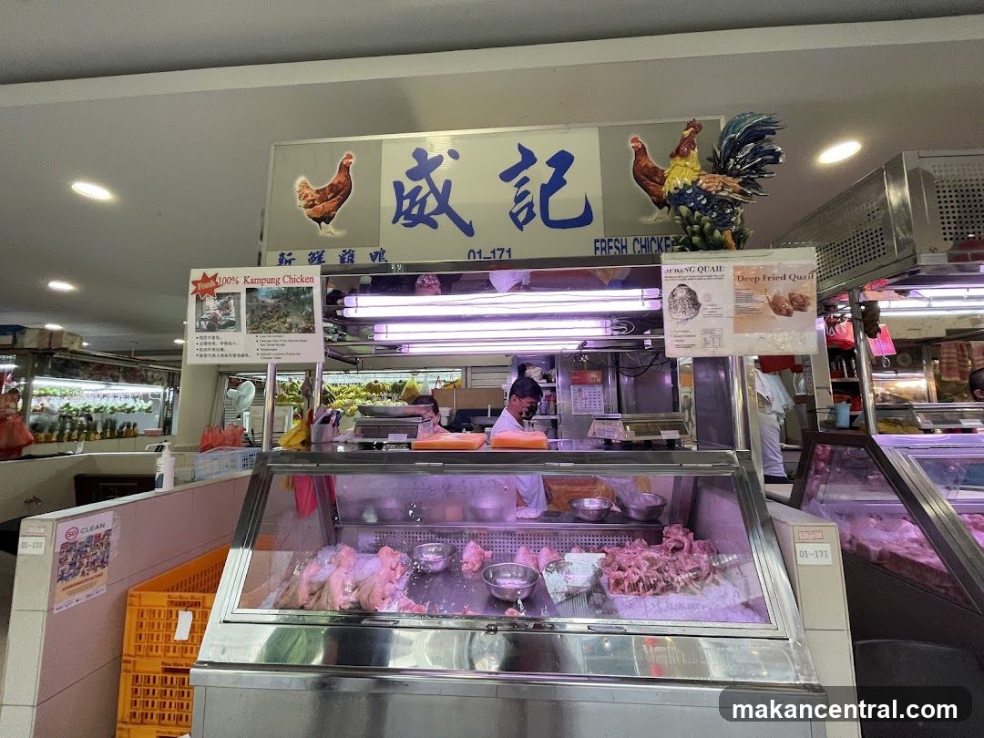 A stall selling fresh chicken and quail. The stall has a large sign with Chinese characters and images of chickens. Below the sign, there is a display case with raw chicken and quail. There are also posters advertising "100% Kampung Chicken" and "Spring Quail". A worker is visible behind the counter.