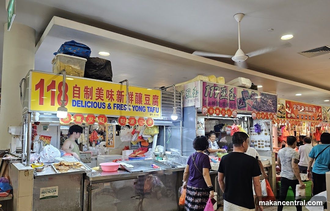 A food stall with a yellow sign that reads "118 自制美味新鮮釀豆商 DELICIOUS & FRES YONG TAFU". There are people ordering and staff preparing food. The stall appears to be in a market or food court setting.