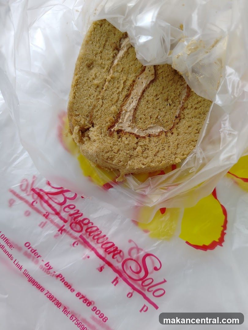 A slice of Swiss roll cake with cream filling, wrapped in plastic. The cake is on a plastic bag with the Bengawan Solo logo and contact information printed on it.