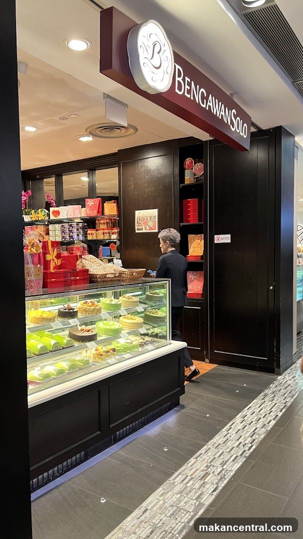 A view of the exterior of a Bengawan Solo bakery. The shop has a display case filled with cakes and pastries. Behind the counter, shelves are stocked with gift boxes and other packaged goods. A person in a dark suit is standing behind the counter. The shop's name and logo are visible on a sign above the entrance.