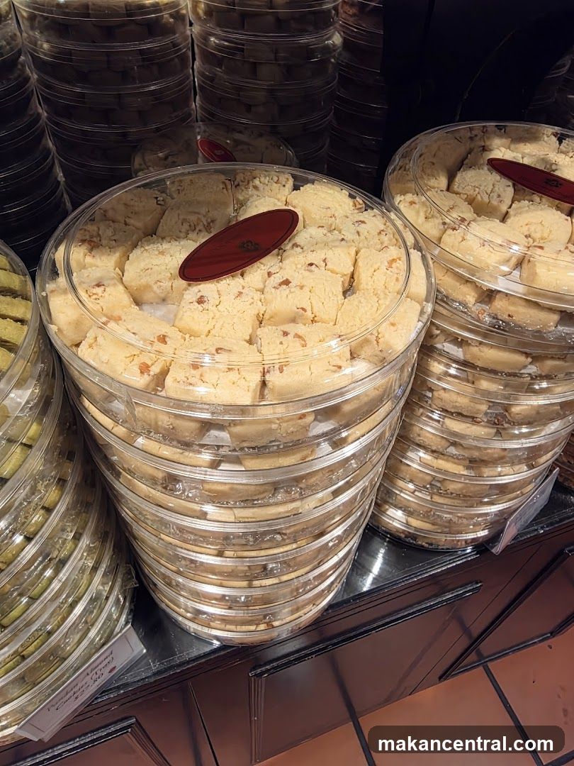 Stacks of clear plastic containers filled with cookies. The cookies in the foreground are light-colored and appear to have nuts or almonds mixed in. A red oval label is affixed to the top container. To the left, another container holds green-colored cookies. A small white tag is visible on the left side, partially obscured, with text that appears to read "Pandan Almond Cookies $22.50".