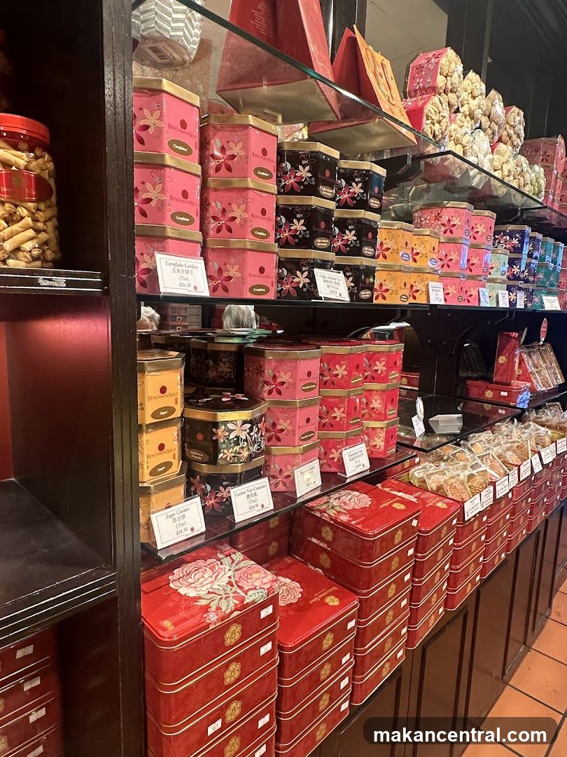 A display shelf filled with various tins of cookies and snacks. The tins are stacked and feature floral and patterned designs. Some labels indicate cookie types and prices. A large jar of rolled cookies is visible on the left.