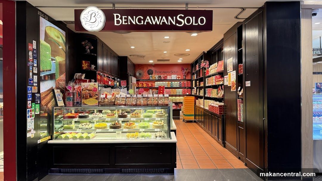 A bakery shop front with a display case full of cakes and pastries. The shop's name, Bengawan Solo, is displayed prominently above the entrance. Shelves behind the counter are stocked with various packaged goods.