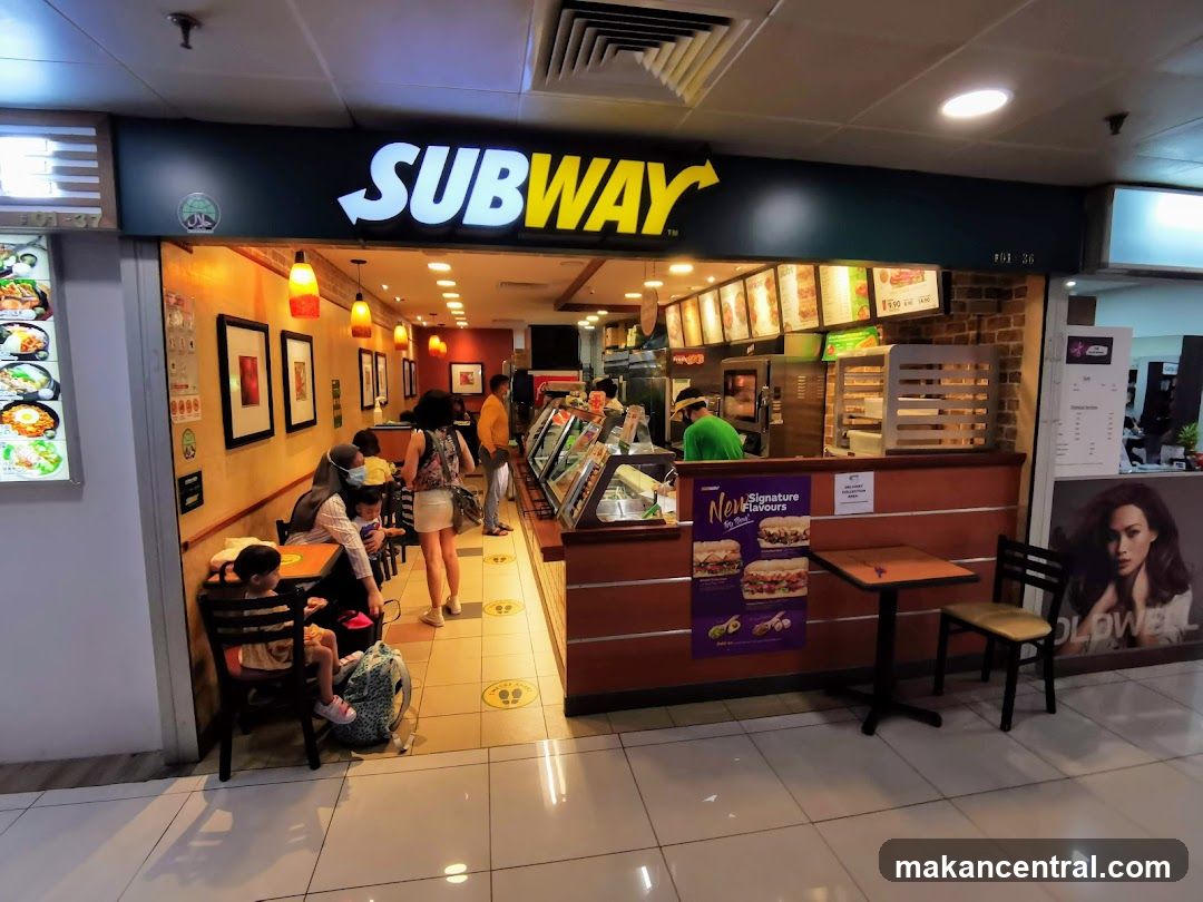 The exterior of a Subway restaurant with its prominent logo above the entrance. Inside, customers are seen dining and ordering. The counter displays various food options, and promotional posters for "Signature Flavours" are visible.