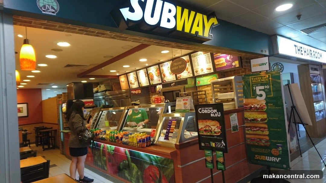 The exterior of a Subway restaurant with a prominent "SUBWAY" sign above the entrance. Inside, a counter displays various food items, and a staff member is visible. Menu boards are mounted on the wall behind the counter. A promotional poster for a value meal is displayed to the right.