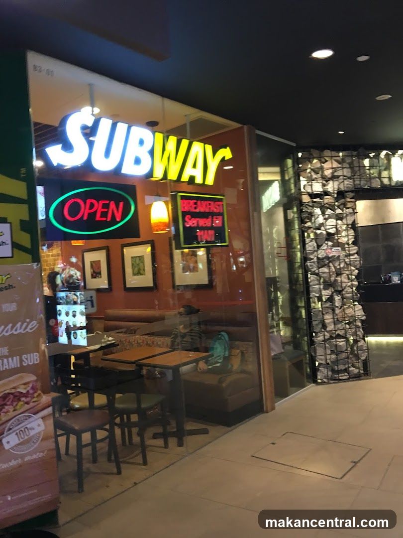 The exterior of a Subway restaurant with a large illuminated sign that says "SUBWAY" and an "OPEN" sign. Inside, tables and seating are visible, along with a person sitting on a bench. A promotional poster for a "Pastrami Sub" is displayed on the left.