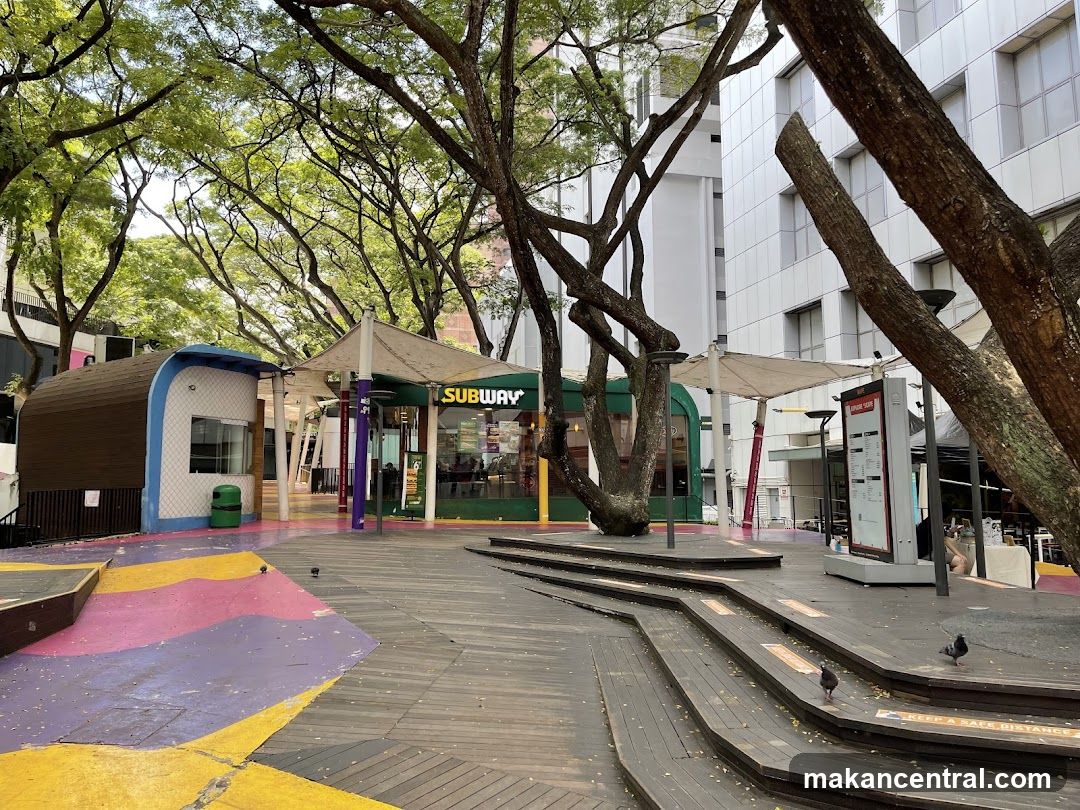 A Subway restaurant storefront is visible with its signature green and yellow signage. The entrance is open, and people can be seen inside. The storefront is situated in an outdoor area with trees and wooden walkways. To the left, there is a small, modern-looking kiosk with a brown and white exterior. In the background, a white building with a grid-like facade rises up. The ground in front of the kiosk and leading towards the Subway is painted with colorful abstract shapes in yellow, purple, and pink. Wooden steps and platforms are also part of the outdoor seating or walkway area.