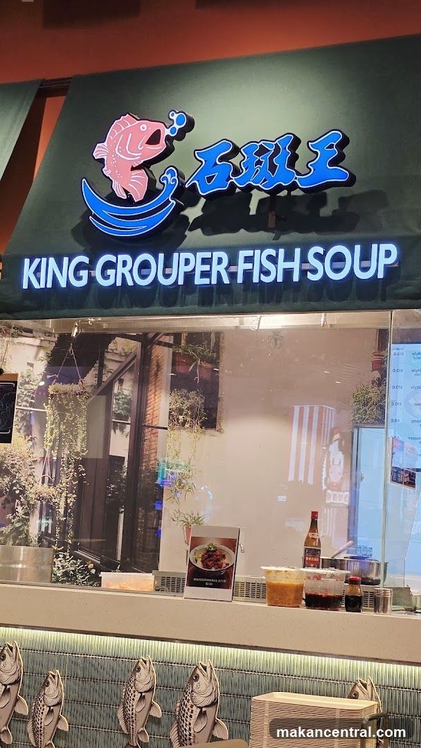 The storefront features a prominent sign with a pink cartoon fish and blue Chinese characters, above the text "KING GROUPER FISH SOUP" in white illuminated letters. Below the sign, a glass partition reveals a display with food items, condiments, and decorative elements. The interior visible through the glass has a backdrop of plants and urban scenery.
