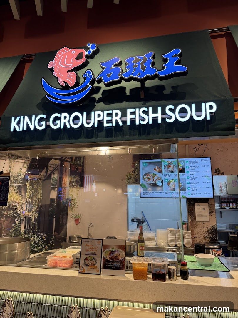 A storefront sign for "KING GROUPER FISH SOUP" with a graphic of a red fish jumping over a blue wave. Below the sign, a glass partition reveals a food stall counter with various condiments, bowls, and a menu board.