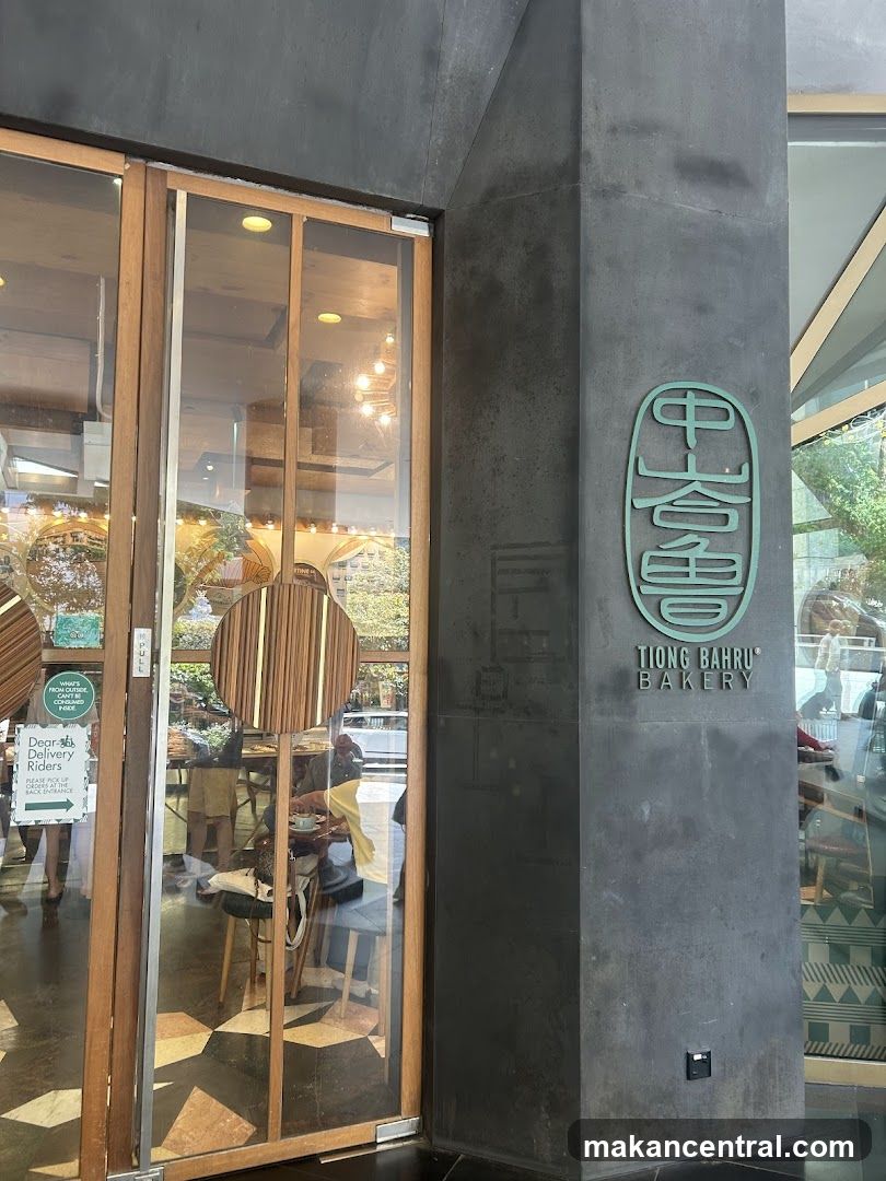 The exterior of a bakery with large glass doors and wooden accents. A dark grey pillar to the right of the entrance features the bakery's logo and name,