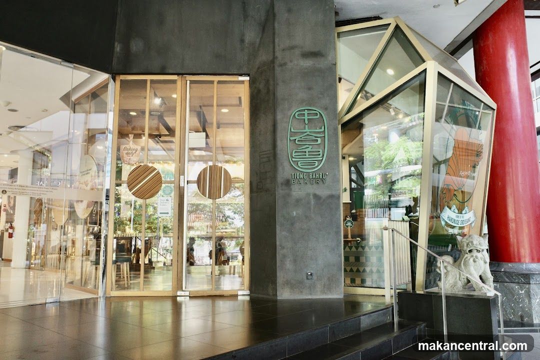The exterior of Tiong Bahru Bakery features a modern design with large glass windows and wooden accents. A prominent green logo with Chinese characters and the text "TIONG BAHRU BAKERY" is visible on a dark grey pillar. The entrance has double glass doors with circular wooden handles. Inside, the bakery appears well-lit with displays of baked goods. A decorative glass structure with an illustration of a croissant is also part of the storefront.
