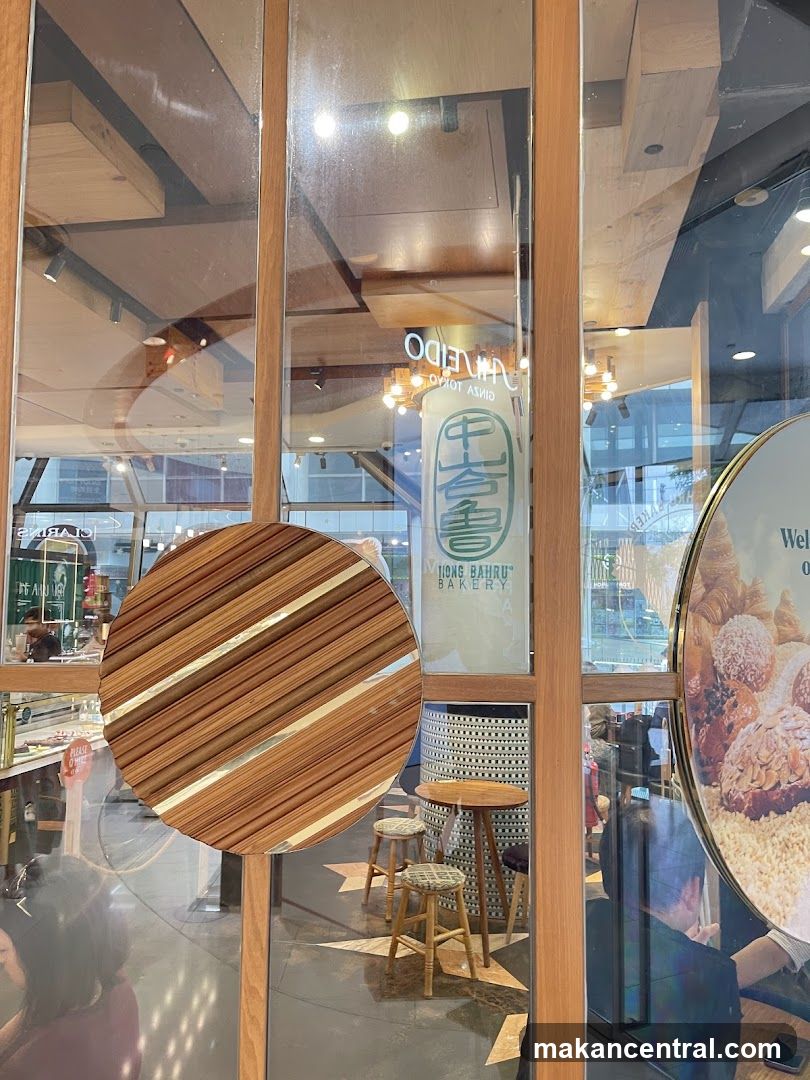 A view through a glass door into a bakery. A circular wooden handle is visible on the door. To the right, a large circular sign displays images of pastries. A vertical sign with green lettering and Chinese characters reads "TIONG BAHRU BAKERY". Inside, there are small tables and stools, and a display case with baked goods.