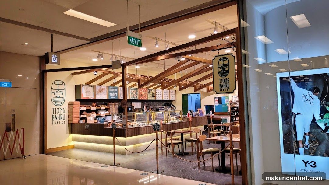 The exterior of Tiong Bahru Bakery, a shop with a warm, inviting ambiance. The bakery features a wooden interior with exposed beams and a display of freshly baked goods. Seating areas with wooden tables and chairs are visible, as well as signage indicating "EXIT" and accessibility information. A large advertisement for Y-3 is displayed on the adjacent storefront.