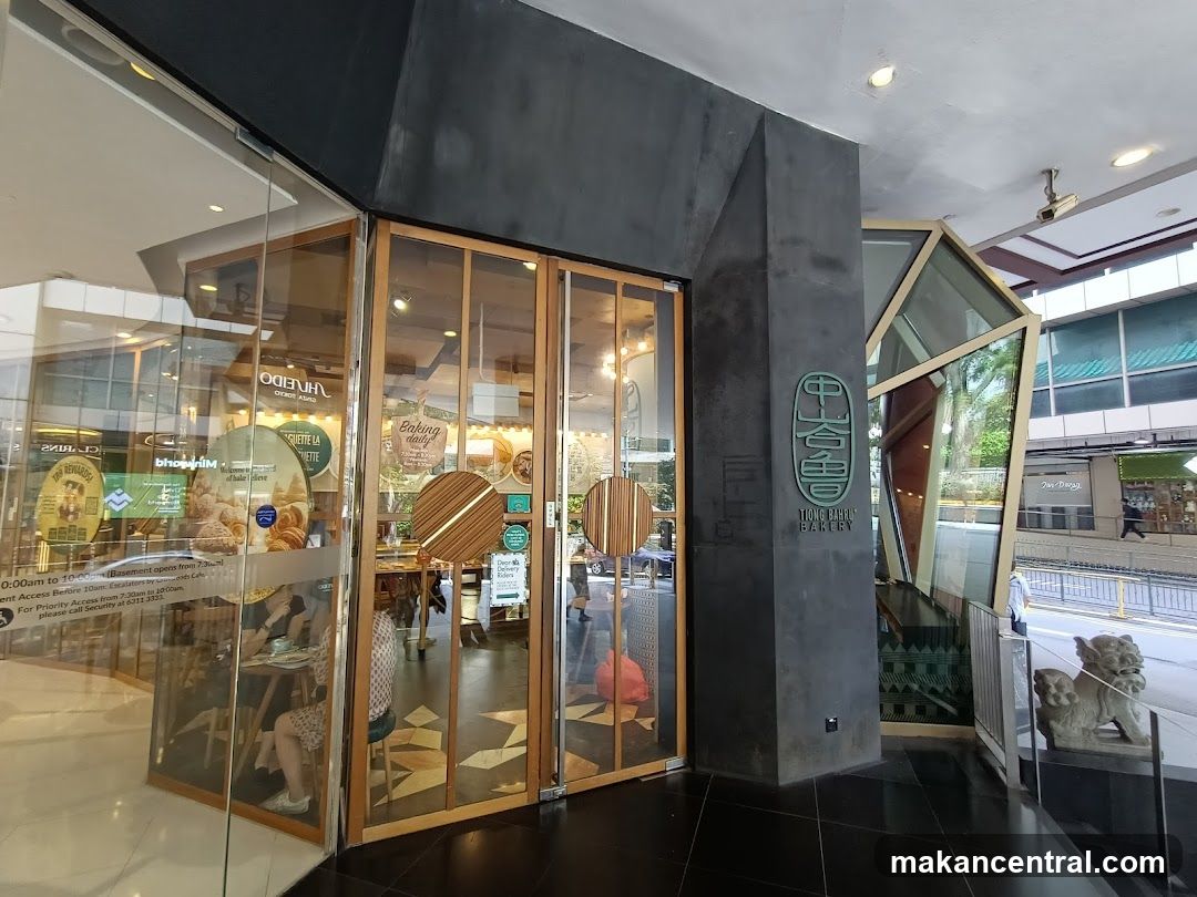 The exterior of a bakery with large glass doors and windows. A sign with Chinese characters and "TIONG BAHRU BAKERY" is visible on a dark grey pillar. Inside, tables and chairs are set up, and baked goods are displayed.