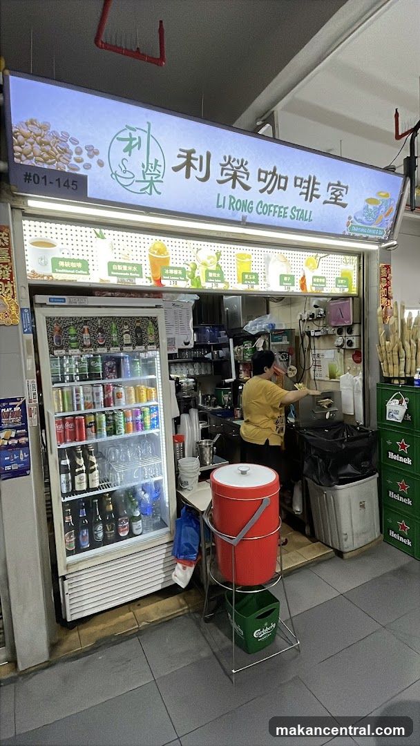 A coffee stall with a bright illuminated sign displaying the name "LI RONG COFFEE STALL" in English and Chinese. Below the sign, a menu board shows various drinks with prices. To the left, a refrigerator is stocked with canned and bottled beverages. A person is seen working behind the counter.