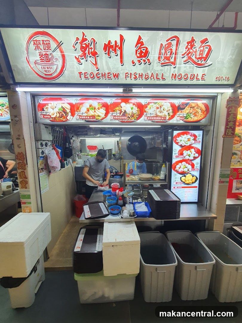 A food stall with a prominent sign displaying "Teochew Fishball Noodle" in English and Chinese. Below the sign are several images of noodle dishes with prices. Two staff members are visible inside the stall, one preparing food and the other serving. In front of the stall are several large containers and stacked boxes.