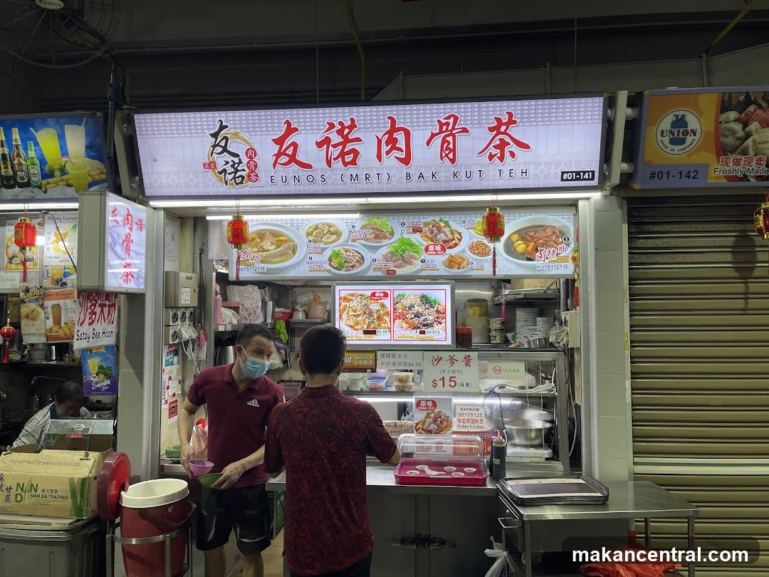 A food stall with a large illuminated sign displaying the name "Eunos (MRT) Bak Kut Teh" in English and Chinese. The sign also features images of various dishes. Two men are visible at the counter, one wearing a mask and holding bowls. Various food items and drinks are displayed on posters and within the stall.