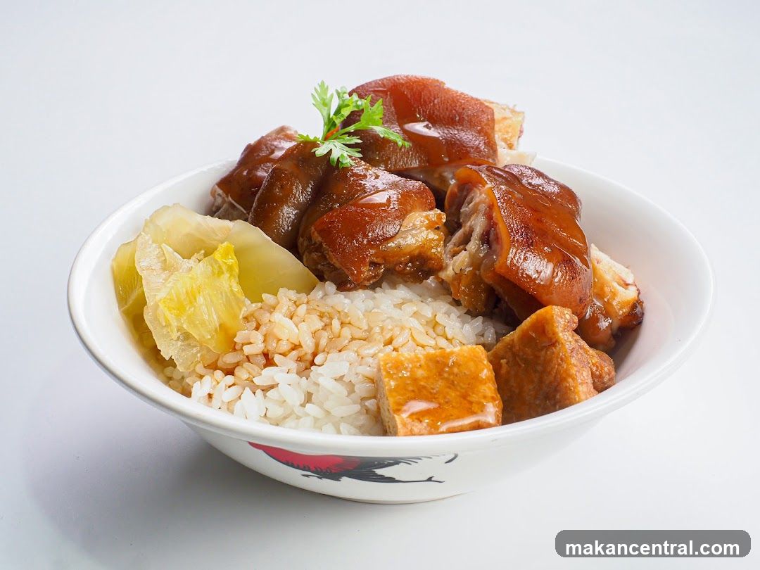 A bowl of rice topped with braised pork knuckles, pickled cabbage, and fried tofu. The pork knuckles are glazed with a dark, savory sauce and garnished with a sprig of cilantro. The rice is fluffy and white, with some sauce drizzled over it. The pickled cabbage is a light yellow-green, and the fried tofu pieces are golden brown. The bowl has a decorative pattern of a rooster on its side.