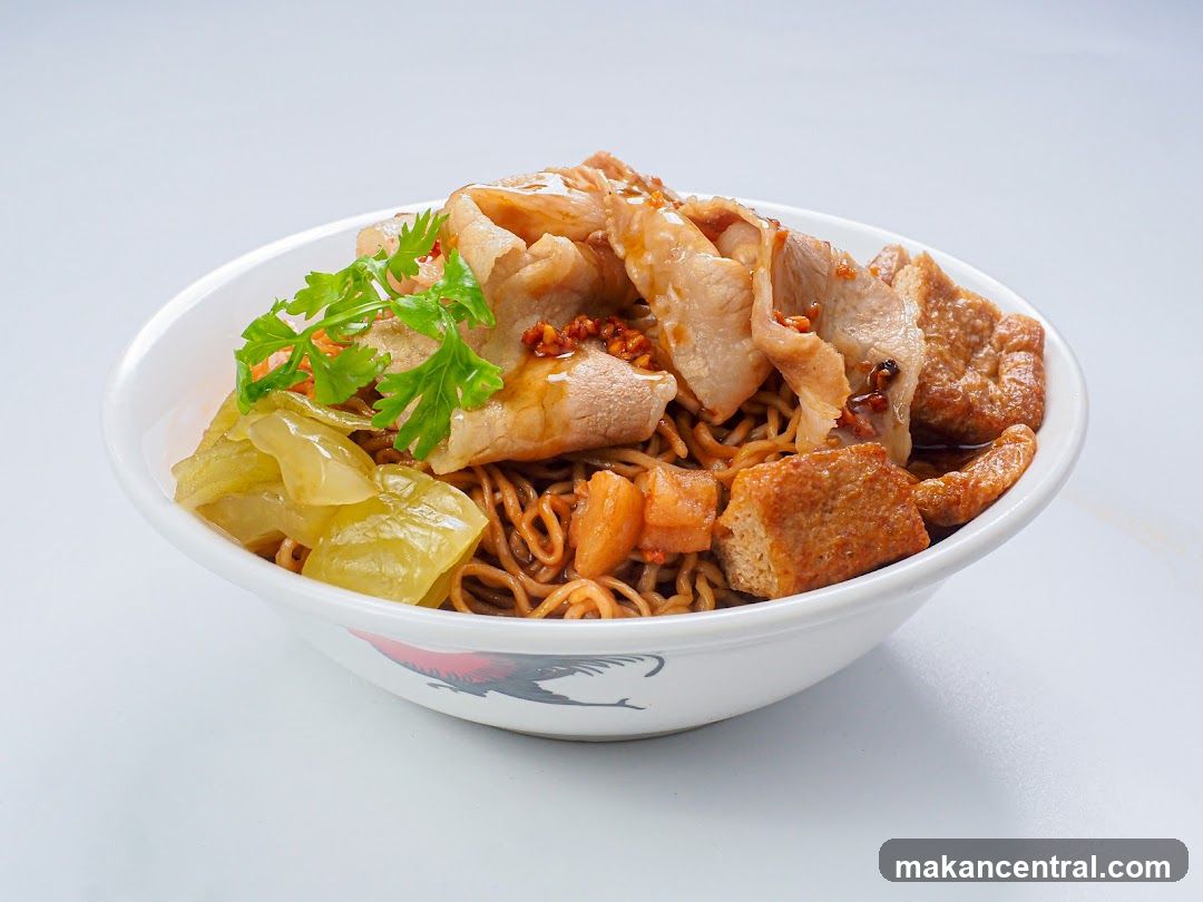 A white bowl filled with noodles, topped with thinly sliced pork, fried tofu cubes, pickled vegetables, and garnished with cilantro. The bowl has a red and black rooster design around its base.