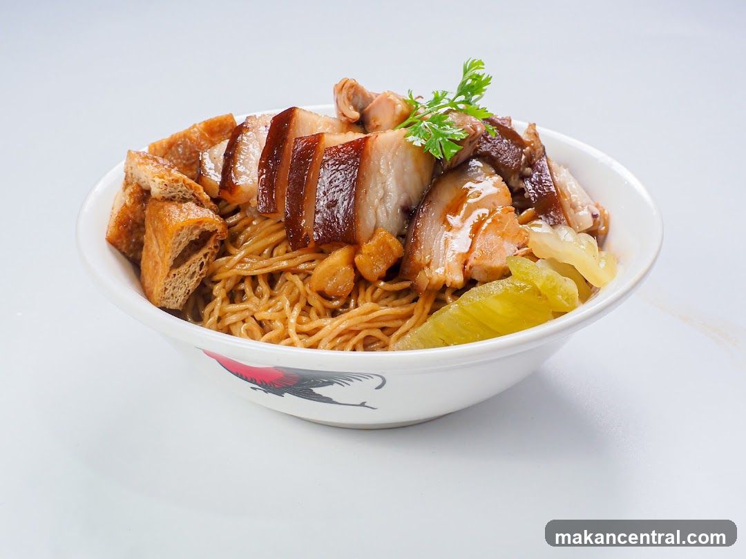 A bowl of noodles topped with braised pork belly, fried tofu, and pickled vegetables, garnished with cilantro. The bowl has a rooster design on its exterior.