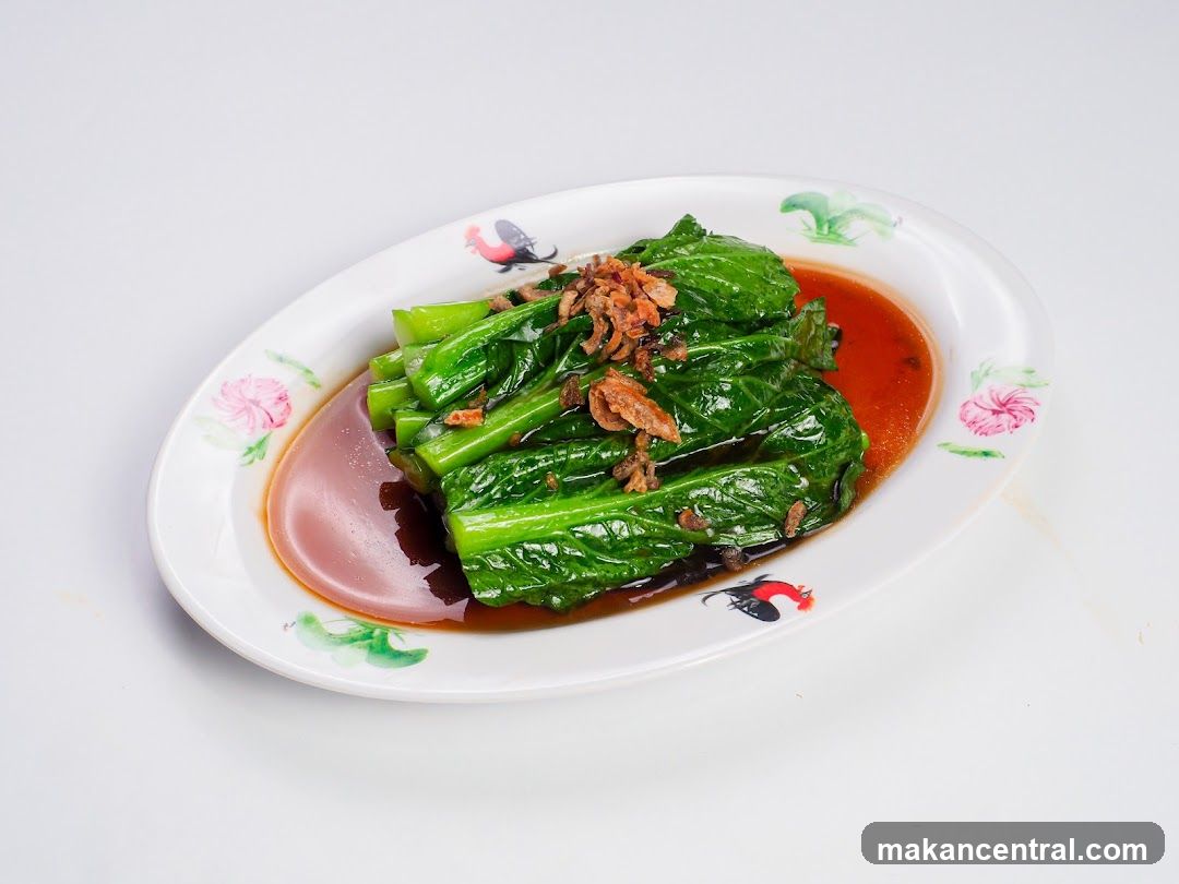 A serving of stir-fried Chinese broccoli, also known as Gai Lan, is presented on a white oval plate with a decorative border featuring floral motifs and a rooster illustration. The vibrant green stalks and leaves of the broccoli are coated in a glossy, savory sauce, and topped with crispy fried shallots. The dish is photographed against a clean white background, highlighting its fresh appearance.
