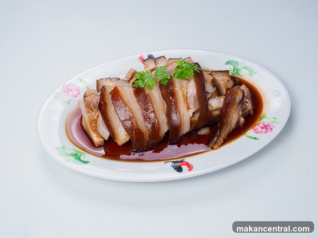 A platter of sliced braised pork belly in a dark sauce, garnished with cilantro, on a white oval plate with floral and rooster designs.