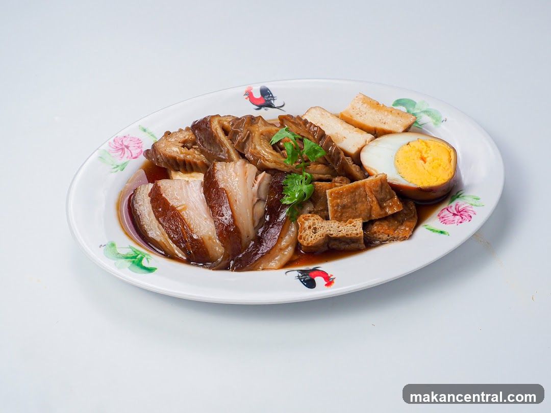 A platter of braised pork belly, sliced and served in a dark sauce. Accompanying the pork are pieces of braised tofu and a halved hard-boiled egg with a bright yellow yolk. The dish is garnished with fresh cilantro and served on a white oval plate decorated with floral patterns and roosters.