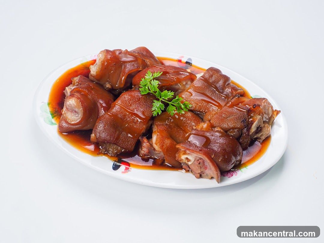 A platter of braised pork knuckles in a rich, dark sauce, garnished with fresh parsley. The pork is tender and glistening, with visible layers of meat and skin. The platter is oval-shaped with a decorative floral rim.