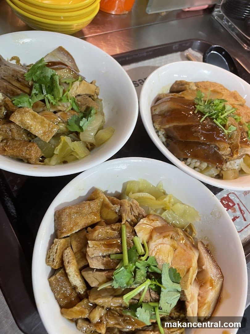 Three white bowls filled with rice and topped with braised meat, tofu, and pickled vegetables. The bowls are arranged on a tray. The meat appears to be pork and duck. The tofu is fried and cut into rectangular pieces. The pickled vegetables are light green and chopped. Fresh cilantro is sprinkled on top of the meat. The lighting is bright, suggesting an indoor setting, possibly a food court or restaurant.