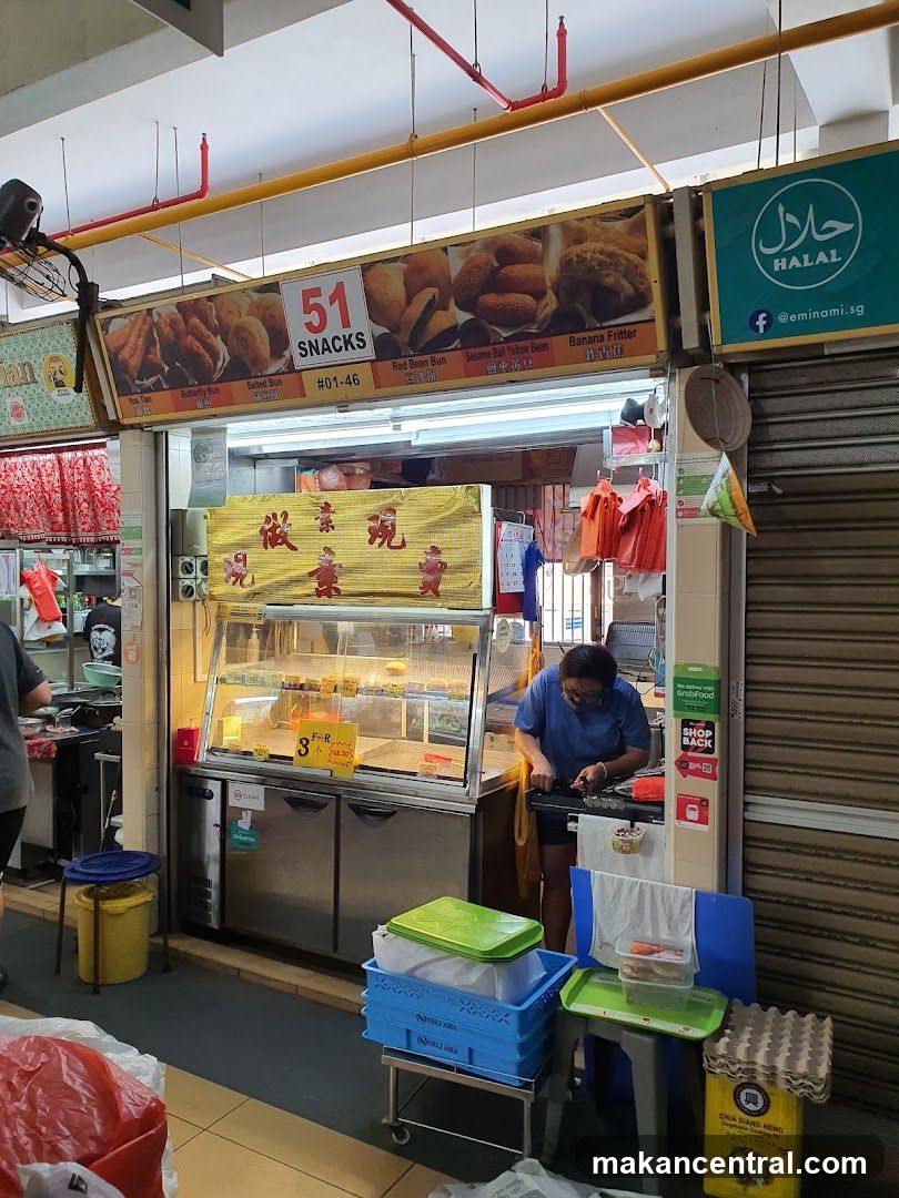 A food stall with a display case filled with snacks. A person is preparing food behind the counter. There is a sign above the stall advertising various snacks like Red Bean Bun, Sesame Ball Yellow Bean, and Banana Fritter. A Halal certification logo is also visible.