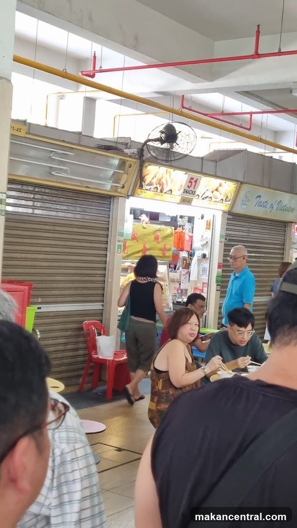 A food stall with a metal roller shutter partially open, revealing a counter and food display. Above the counter, a sign displays "51 SNACKS" and images of food. To the right, a sign reads "Taste of Vietnam". People are seated at tables in front of the stall, eating.