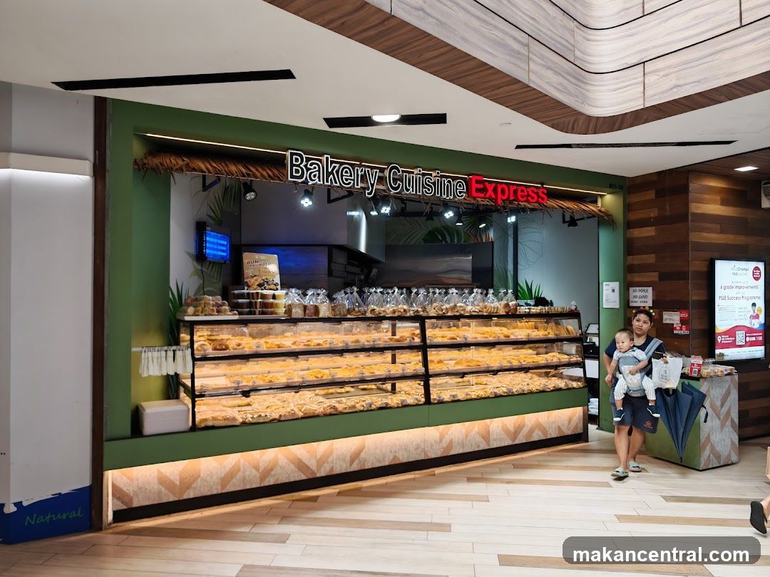 A bakery storefront with a display case full of pastries and bread. A woman is standing to the right of the display case holding a baby. There is a promotional poster on the wall to the right of the woman.