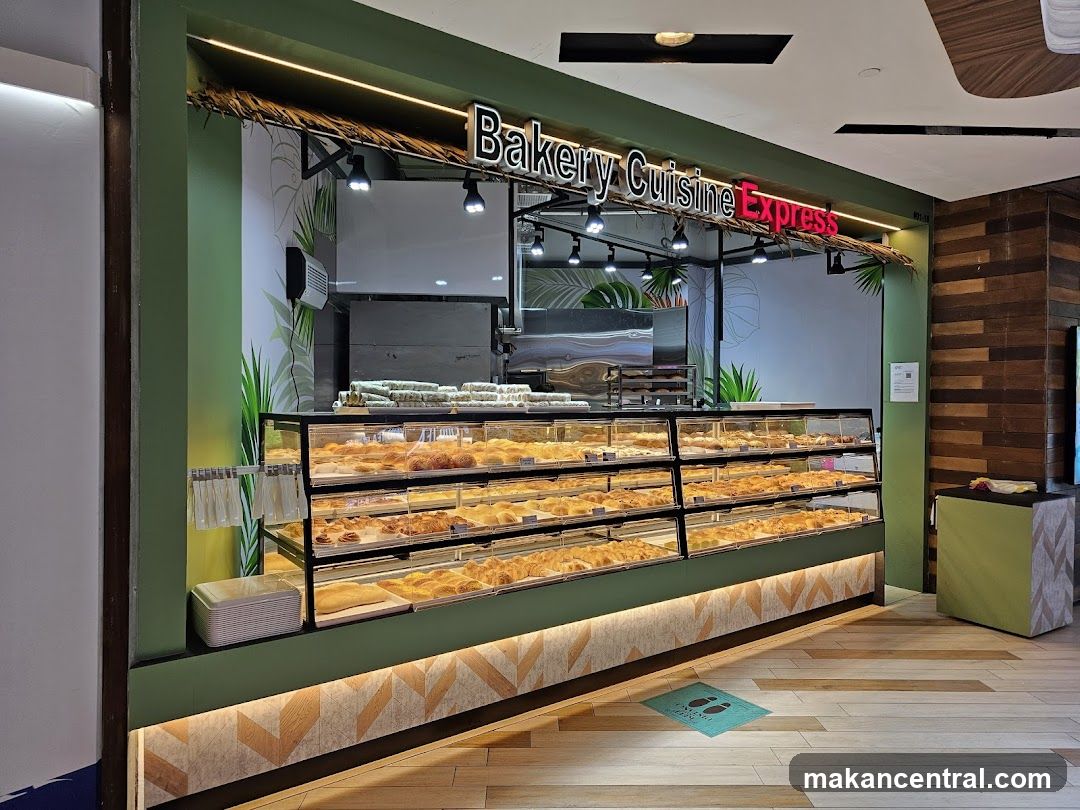 A bakery display case filled with various pastries and baked goods. The storefront has a green facade with a thatched roof overhang. The sign above reads "Bakery Cuisine Express" in illuminated letters. Tropical leaf motifs are painted on the back wall.