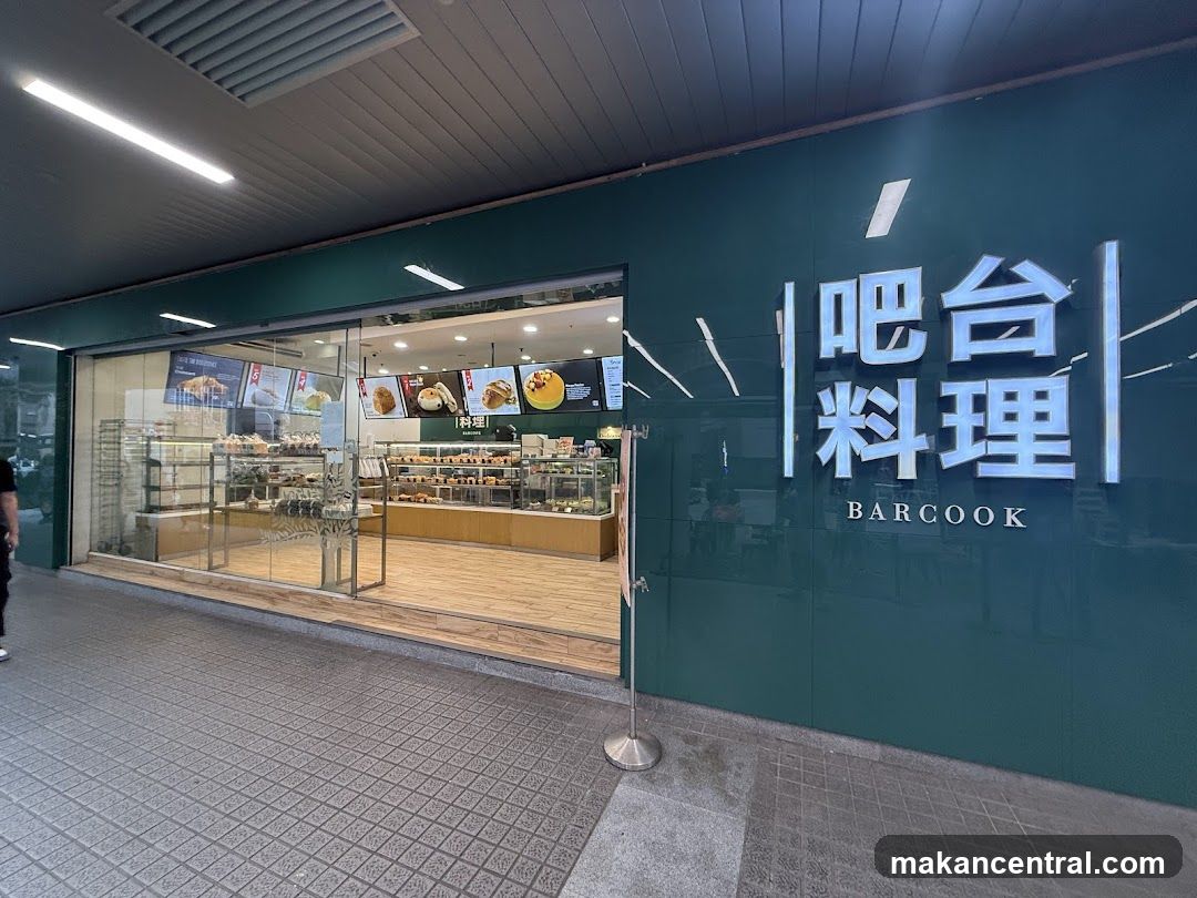 The exterior of a Barcook bakery with a large green facade featuring the store's name in illuminated Chinese characters and English text 'BARCOOK'. The entrance is a glass storefront revealing shelves stocked with baked goods and display cases. Inside, more pastries and breads are visible on shelves and in display cases. Digital menu boards are mounted above the counter inside the shop.