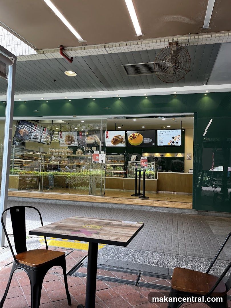 The exterior of a bakery with a green facade and large glass windows displaying baked goods. There is an outdoor seating area with a table and chairs in front of the shop. Inside, shelves are filled with bread and pastries. Digital screens display images of food and text.