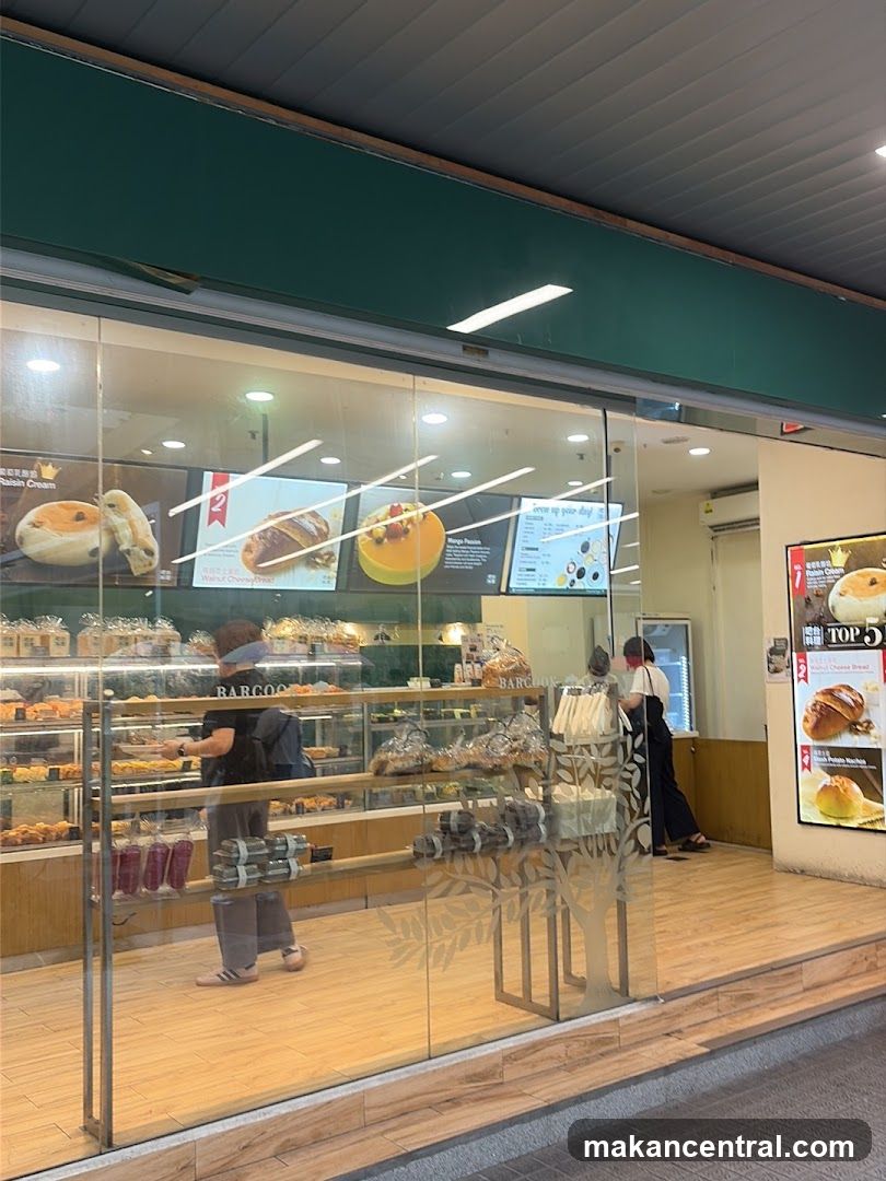 A view of a bakery's storefront, showcasing a display of baked goods through large glass windows. People are seen inside browsing the shelves. The interior features wooden flooring and glass display cases filled with various breads and pastries. A menu board is visible inside, along with promotional images of food items. The shop's name,